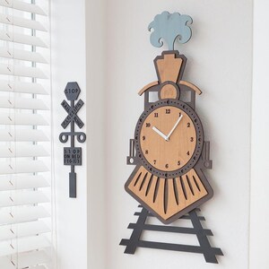 Può includere: Orologio da parete in legno a forma di treno con quadrante marrone chiaro e cornice marrone scuro. L'orologio ha lancette nere e una cornice esterna marrone scuro. È visibile anche un segnale di passaggio a livello in legno abbinato. L'orologio è alto circa 51 cm.