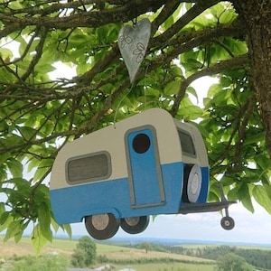 Puede incluir: Un original comedero para pájaros hecho a mano con forma de caravana vintage. El comedero está pintado en crema y azul, con ruedas negras y una pequeña puerta. Un adorno metálico en forma de corazón cuelga encima.