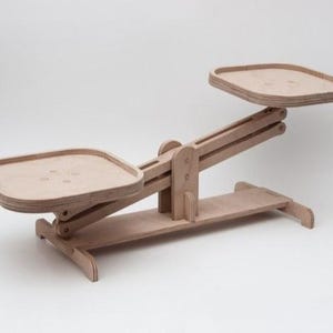 May include: A wooden balance scale with two square trays and a pivoting arm. The scale is made of light-colored wood and has a simple, geometric design. The trays are approximately 10 inches square.