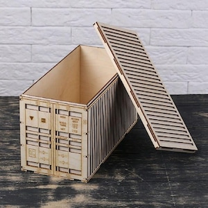 Puede incluir: Una caja de madera con forma de contenedor de envío, con tapa con bisagras. La madera clara presenta listones verticales en los laterales y detalles en el frente. La tapa está parcialmente abierta, revelando el interior.
