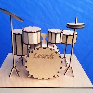 Op de afbeelding: Een miniatuur houten drumstel met een natuurlijke houten afwerking en zwarte accenten. De set bevat een basdrum met de naam "Leerok", twee toms, een snaredrum en bekkens. De drums zijn op een houten basis gemonteerd.