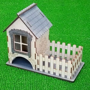 May include: A wooden miniature house with a gray shingled roof, a decorative window frame, and a white picket fence. The house is made of light-colored wood and sits on a gray base. The background is green.