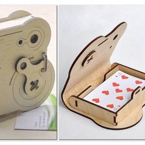 Può includere: Scatola di carte da gioco in legno dal design unico. La scatola è marrone chiaro e ha una chiusura a scatto. La scatola aperta rivela una carta da gioco con cuori rossi. Il design ricorda una fotocamera vintage.