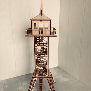 以下が含まれることがあります： 尖った屋根と、窓とバルコニーのある小さな建物が上部にある高い塔の木製模型。塔は格子状の構造で、三脚ベースで支えられています。模型は明るい茶色の木材で作られています。