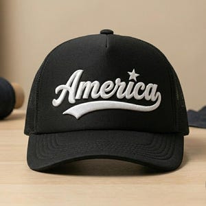 Puede incluir: Una gorra tipo trucker negra con un panel frontal estructurado que presenta la palabra America bordada en letras cursivas blancas. Una pequeña estrella blanca está situada sobre la letra i. La gorra incluye una visera curva y paneles laterales de malla.
