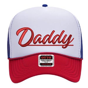 May include: A red, white, and blue trucker hat with the word "Daddy" embroidered in red script. The hat has a white foam front, a red curved peak, and a blue mesh back. The hat is a classic style.