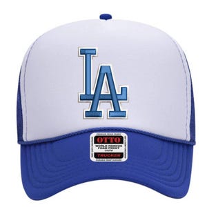 May include: A blue and white trucker cap featuring the letters "LA" in blue. The cap has a blue peak and a white foam front. It includes a snap closure and an "OTTO" tag. The design is suitable for casual wear.