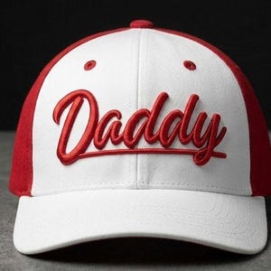 Può includere: Un cappellino da baseball bianco con visiera rossa e la scritta "Daddy" ricamata in rosso sul davanti. Il cappellino ha una visiera curva e un bottone sulla parte superiore.