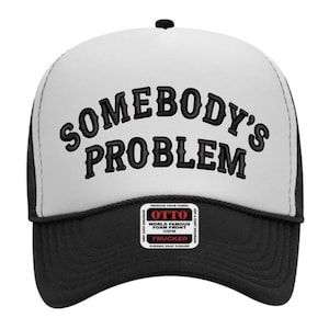 May include: A black and white trucker hat with the embroidered text "SOMEBODY'S PROBLEM" in a bold, stylized font. The hat features a white front panel, a black brim, and a black mesh back. A small tag is visible on the front.