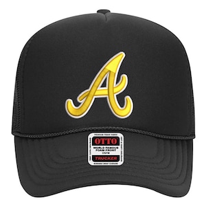 Könnte beinhalten: Schwarze Trucker-Mütze mit gelbem gesticktem "A"-Logo. Die Mütze hat eine Schaumstofffront und einen Mesh-Rücken. Die Mütze hat einen Schnappverschluss und den Text "OTTO WORLD FAMOUS FOAM FRONT OSFM TRUCKER" auf einem kleinen Etikett.