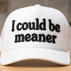 Könnte beinhalten: Weißer Trucker-Cap mit dem gestickten Schriftzug "I could be meaner" in schwarzen Blockbuchstaben. Die Kappe hat eine Mesh-Rückseite und einen gebogenen Schirm. Ein lässiges Accessoire.