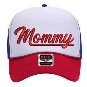 May include: A red, white, and blue trucker hat with the word "Mommy" embroidered in red script across the front. The hat features a white foam front, a red brim, and a blue mesh back.