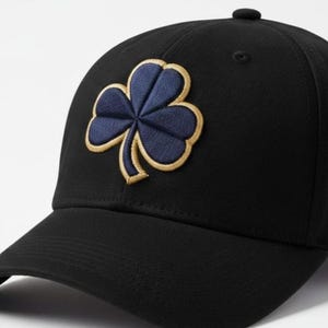 Può includere: Cappellino da baseball nero con un trifoglio ricamato blu navy e oro. Il cappellino ha una visiera curva e un bottone sulla parte superiore. Un accessorio alla moda per l'uso quotidiano.