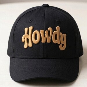 Pode incluir: Boné de beisebol preto com a palavra "Howdy" bordada em uma fonte estilizada marrom na frente. O boné tem uma aba curva e um botão na parte superior. Um acessório casual.