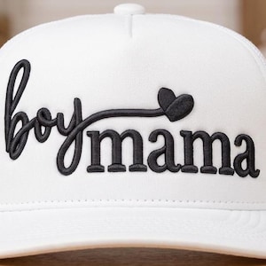 Pode incluir: Boné de caminhoneiro branco com as palavras "boy mama" bordadas em preto, em letras cursivas e maiúsculas, com um coração. O boné tem uma parte traseira de malha e uma viseira curva.