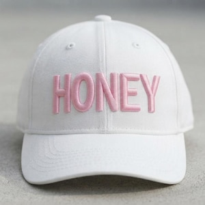 Può includere: Un cappellino da baseball bianco con la parola HONEY ricamata in lettere rosa chiaro in rilievo sul pannello frontale. Il cappellino presenta una corona strutturata, una visiera curva e occhielli bianchi abbinati sui pannelli superiori.