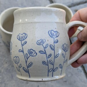 May include: A ceramic mug with a speckled cream-colored glaze featuring a hand-drawn blue floral pattern of stems and petals. The mug has a rounded body, a wide rim, and a sturdy side handle. The design includes multiple flower stalks.