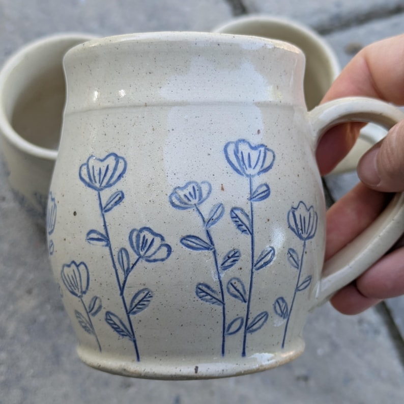 May include: A ceramic mug with a speckled cream-colored glaze featuring a hand-drawn blue floral pattern of stems and petals. The mug has a rounded body, a wide rim, and a sturdy side handle. The design includes multiple flower stalks.