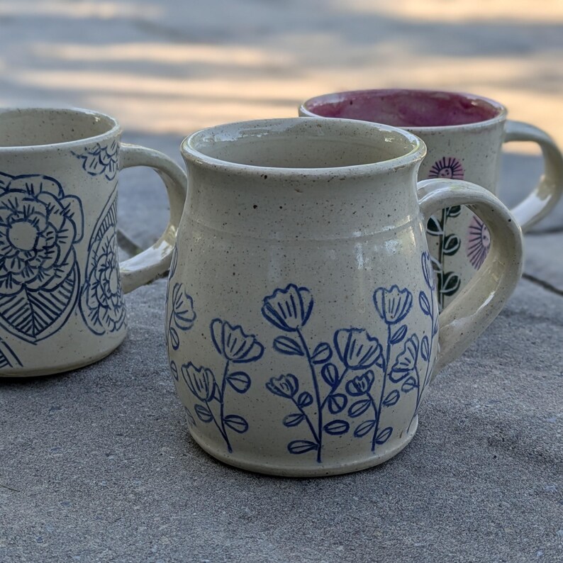 May include: Three ceramic mugs with a speckled cream finish. The front mug features blue line-art flowers. The left mug displays a large blue floral pattern. The back mug has a pink interior and a purple flower design on the exterior surface.