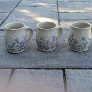 May include: Three ceramic mugs with a cream-colored base and a blue hand-painted floral pattern. Each mug features a rounded body, a sturdy handle on the side, and a smooth rim. The blue floral design wraps around the lower portion of each mug.