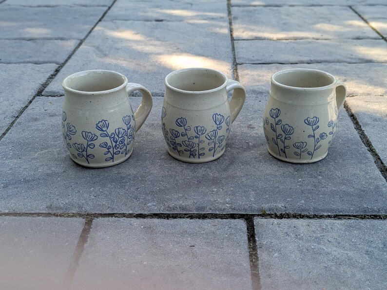 May include: Three ceramic mugs with a cream-colored base and a blue hand-painted floral pattern. Each mug features a rounded body, a sturdy handle on the side, and a smooth rim. The blue floral design wraps around the lower portion of each mug.