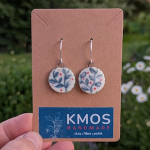 May include: A pair of round, white ceramic drop earrings featuring a hand-painted blue leaf pattern with small red dots. The earrings hang from silver-toned metal hooks and are displayed on a brown cardboard card labeled KMOS HANDMADE.
