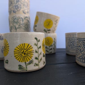 May include: A collection of ceramic cups with floral designs. Some cups feature yellow and black dandelion illustrations, while others display blue floral patterns. The cups are arranged on a dark surface against a light blue backdrop.