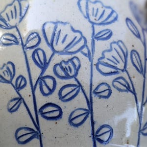 May include: A ceramic surface featuring a hand-painted blue floral pattern on a speckled off-white background. The design consists of multiple vertical stems with simple leaves and stylized flower blossoms, creating a repeating botanical motif.