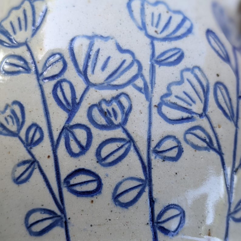 May include: A ceramic surface featuring a hand-painted blue floral pattern on a speckled off-white background. The design consists of multiple vertical stems with simple leaves and stylized flower blossoms, creating a repeating botanical motif.