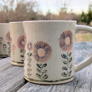 May include: A collection of handmade ceramic mugs with floral designs. The mugs feature a beige base color with blue and pink floral patterns. Some mugs have handles, while others are cylindrical.