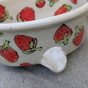 May include: A ceramic bowl with a speckled off-white glaze featuring hand-painted red strawberries with green leaves and black outlines. The bowl has a rounded rim and a small, protruding ceramic handle on the side, showing a glossy finish.