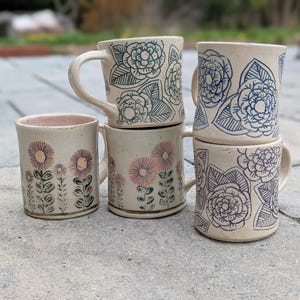 May include: A collection of handmade ceramic mugs with floral designs. The mugs feature a beige base color with blue and pink floral patterns. Some mugs have handles, while others are cylindrical.