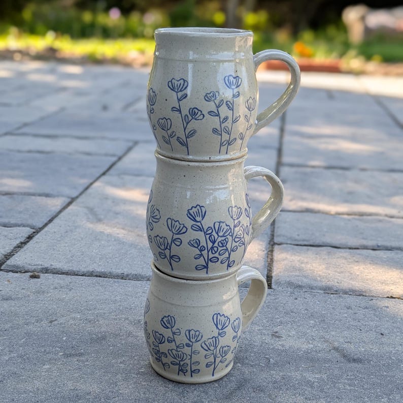 May include: A vertical stack of three ceramic mugs with a speckled cream-colored glaze. Each mug features a hand-drawn blue floral pattern consisting of stems, leaves, and small blossoms. The mugs have a rounded body shape and a single side handle.