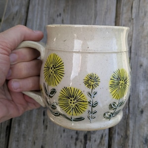 Könnte beinhalten: Cremefarbene Keramiktasse mit gelbem Löwenzahn-Blumendesign. Die Tasse hat eine abgerundete Form mit Henkel und ist mit schwarzen und grünen Akzenten verziert. Die Tasse wird von einer Hand gehalten.
