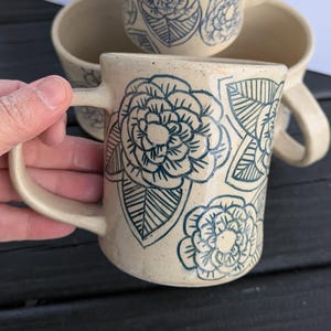 May include: Cream-colored ceramic mug with yellow dandelion flower design. The mug has a rounded shape with a handle and is decorated with black and green accents. The mug is held by a hand.