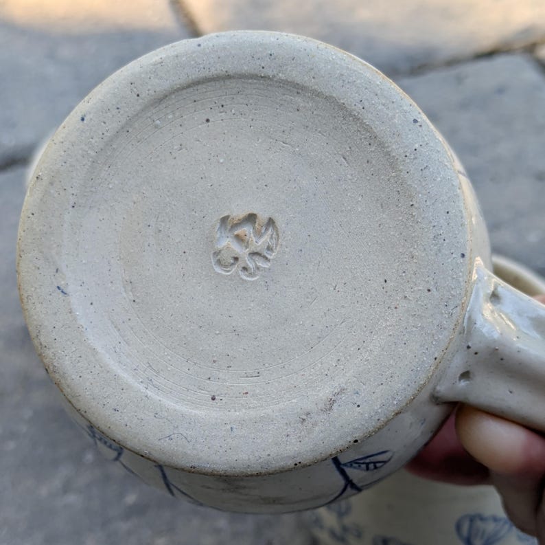 May include: The underside of a speckled beige ceramic mug featuring a circular base with a textured, matte finish. A small, stamped maker's mark is centered on the bottom. The mug has a sturdy handle attached to the side and blue floral patterns.
