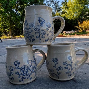 May include: Three ceramic mugs with a speckled cream-colored glaze and a hand-painted blue floral pattern. Each mug features a rounded body, a wide rim, and a sturdy side handle. The floral design consists of simple line-drawn flowers on stems.