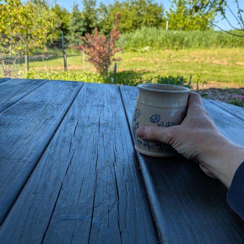 May include: A speckled off-white ceramic mug featuring a hand-painted blue floral pattern with thin stems and petals. The mug has a slightly flared rim and a smooth, rounded body, held against a dark wooden surface with visible grain texture.