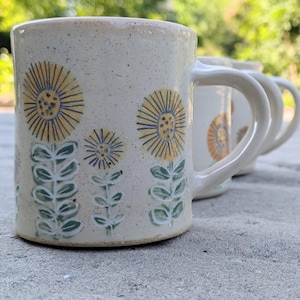 Op de afbeelding: Een keramische mok met een gespikkelde crèmekleurige glazuurlaag, voorzien van handgeschilderde gele bloemen met blauwe harten en groene stengels met bladeren. De mok heeft een gebogen handvat en maakt deel uit van een set van drie mokken.