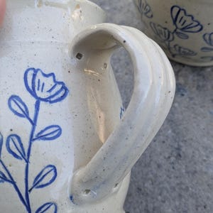 May include: A cream-colored ceramic pitcher with a speckled finish and a blue hand-painted floral design featuring a stem with leaves and a flower. The pitcher has a curved handle attached to the side, and a second matching ceramic piece is visible.