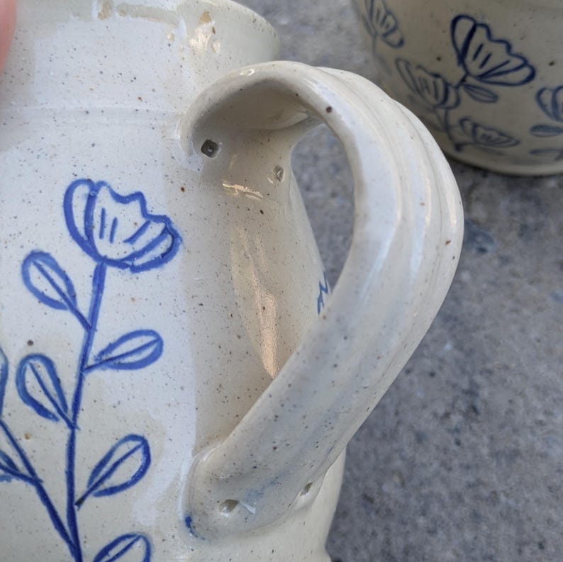 May include: A cream-colored ceramic pitcher with a speckled finish and a blue hand-painted floral design featuring a stem with leaves and a flower. The pitcher has a curved handle attached to the side, and a second matching ceramic piece is visible.