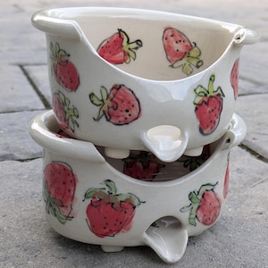 Può includere: Due ciotole in ceramica impilate con smalto color crema e illustrazioni di fragole rosse dipinte a mano con foglie verdi. Ogni ciotola presenta un'ampia apertura curva lungo il bordo e un piccolo beccuccio sporgente alla base.