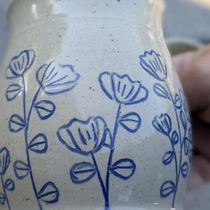 May include: A speckled off-white ceramic mug featuring a hand-drawn blue floral pattern. The design consists of multiple stems with simple petals and leaves etched or painted onto the exterior surface of the cylindrical vessel.