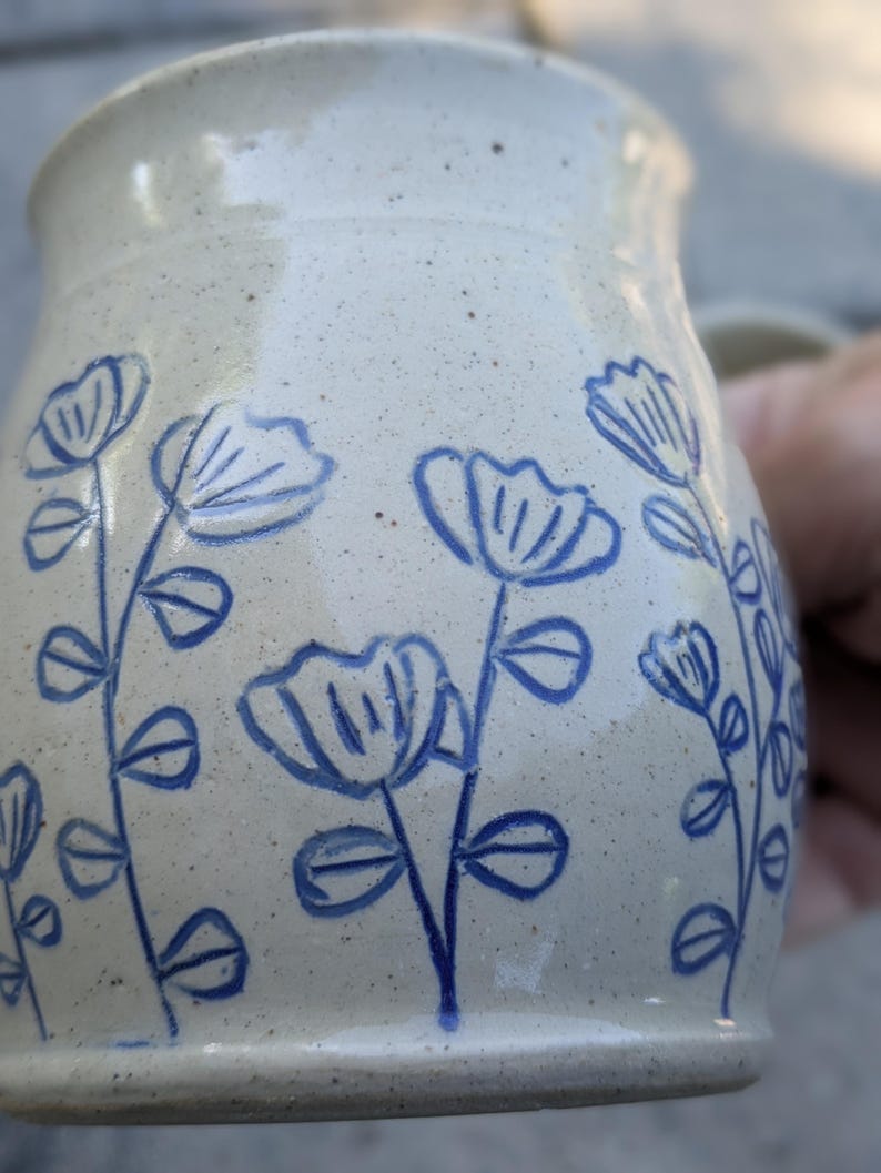 May include: A speckled off-white ceramic mug featuring a hand-drawn blue floral pattern. The design consists of multiple stems with simple petals and leaves etched or painted onto the exterior surface of the cylindrical vessel.