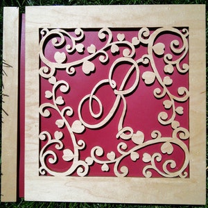 May include: A wooden cutout of a decorative design with a large letter "P" in the centre. The design is made up of swirling vines and hearts, and the background is a deep red colour.