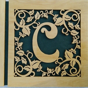 May include: A wooden plaque with a decorative cutout of a leafy vine design surrounding the letter "C". The letter is cut out of the wood and reveals a dark green background.