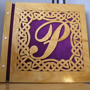 May include: A wooden book cover featuring a Celtic knot design and a carved letter "P" in the centre. The letter is cut out of the wood, revealing a purple velvet background.