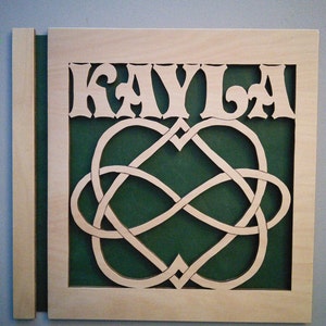 May include: A wooden sign with the name "KAYLA" cut out in a decorative font. The sign features a green background with a Celtic knot design in the shape of two intertwined hearts.