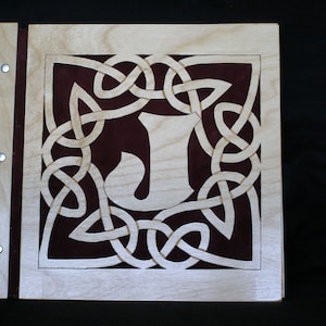 May include: A wooden scrapbook cover featuring a Celtic knot design with the letter "J" at its centre. The knotwork is cut out of the wood, revealing a burgundy background.