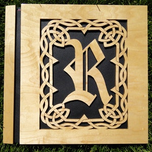 May include: A wooden plaque with a Celtic knot border and a black leather background. The letter "R" is cut out of the wood and placed on the black leather background.